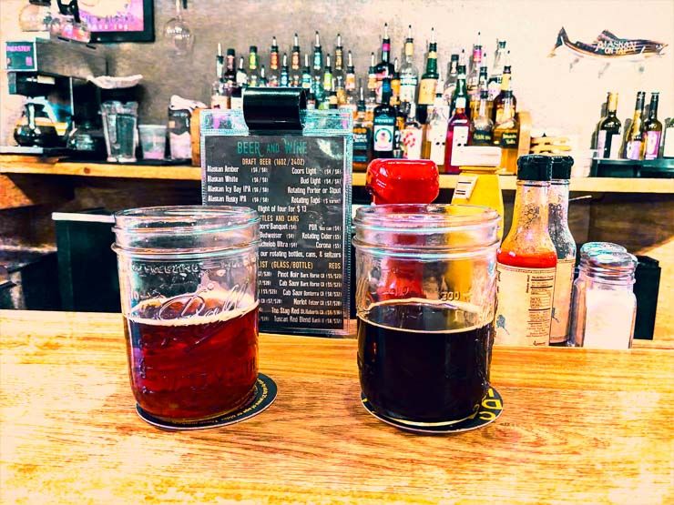 two beers (a red amber ale and a dark stout) on a wooden bar counter top, in front of a beer and wine menu, condiments, and a wall of wine and liquor bottles